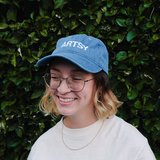 Person wearing a blue cap with 'ARTSY' text against a green leafy background Artsy Embroidered Hat