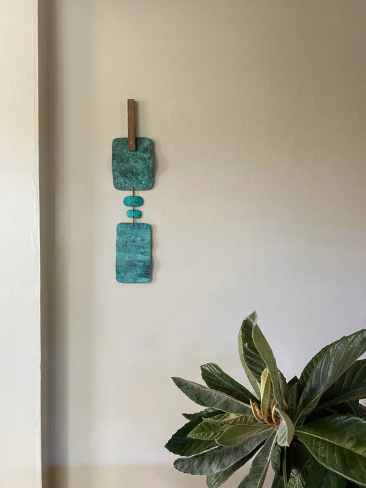 Decorative wall hanging with turquoise square elements on a plain wall. Wall hanging made of patinated brass pieces and hammered brass rod connection and a wooden shape. Handmade in NorCal.