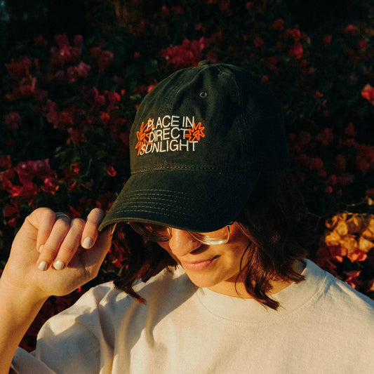 Person wearing a black cap with text and flowers in the background Place in Direct Sunlight Embroidered Hat