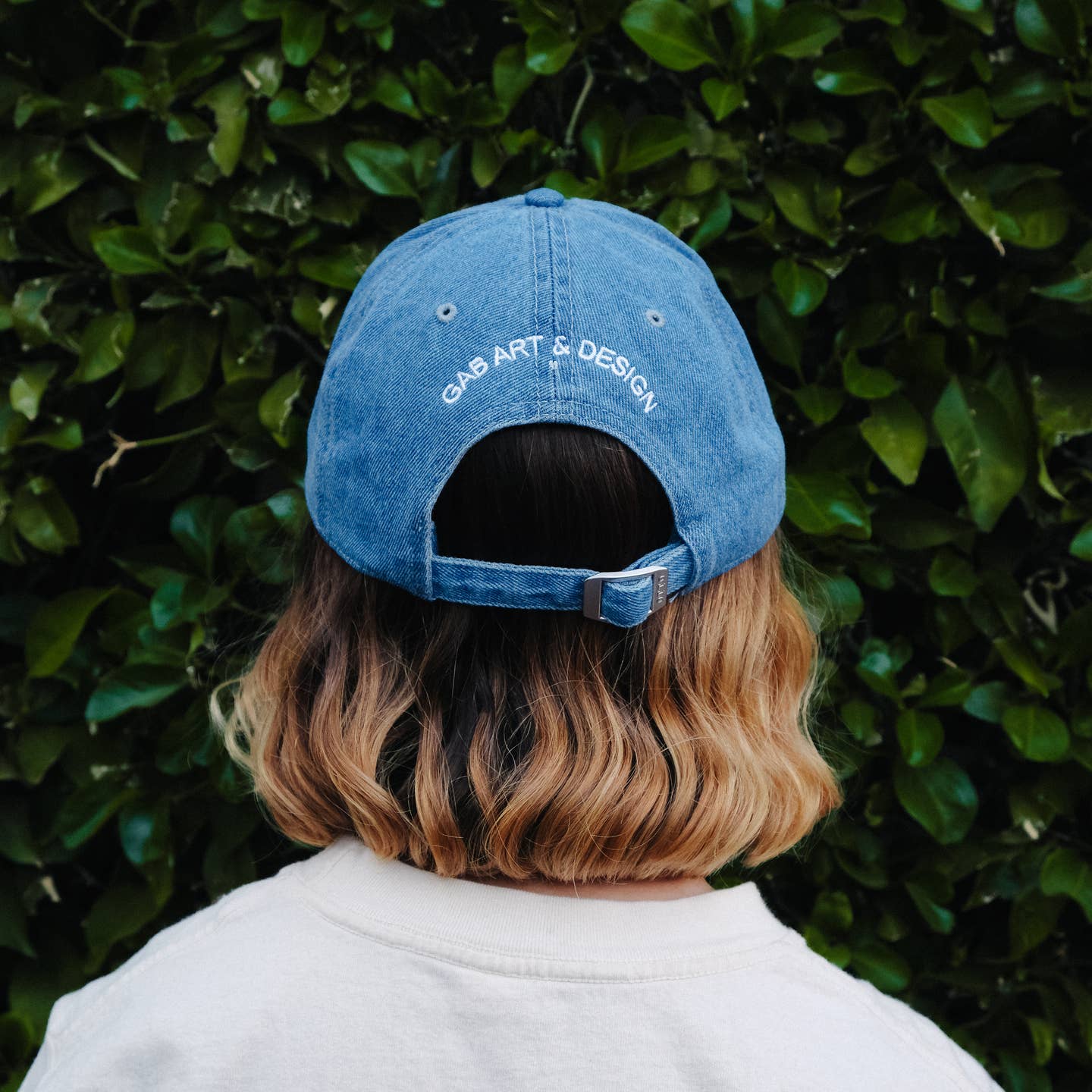 Person wearing a blue cap with 'GAR ART & DESIGN' text against a green hedge background Artsy Embroidered Hat