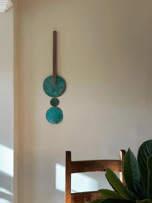 Decorative wall art with circular designs on a beige wall, with a wooden chair and plant in the foreground. Wall hanging made of patinated brass pieces and hammered brass rod connection and a wooden shape. Handmade in NorCal.
