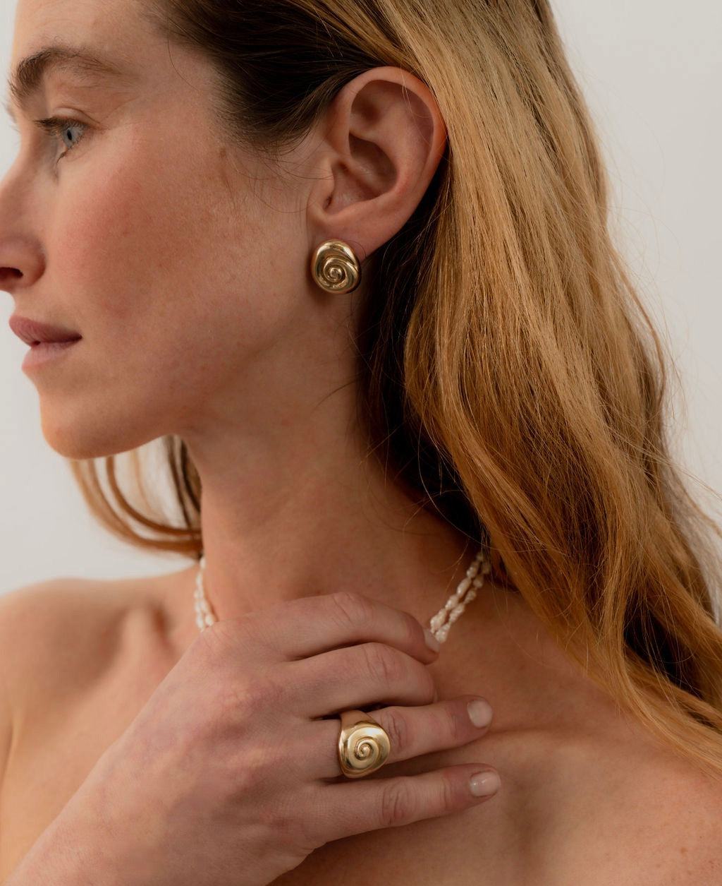 Woman wearing gold spiral earrings and a ring, with a plain background. Amanda Hunt Compass Earrings