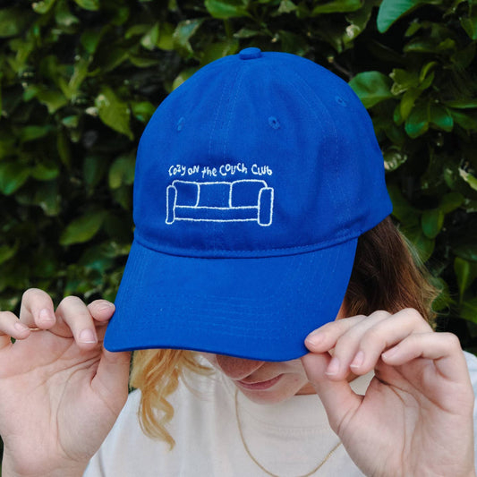 Person holding a blue cap with a logo and text against a green leafy background Cozy On the Couch Club Embroidered Hat