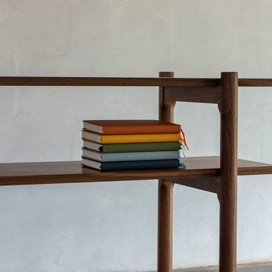 Hardcover paper journals by Anecdote in a variety of colors stacked on a bookshelf.