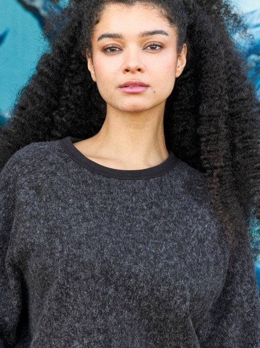 Woman wearing a dark gray sweater with a blurred blue and white background. Mollusk Surf Shop Softest Crew