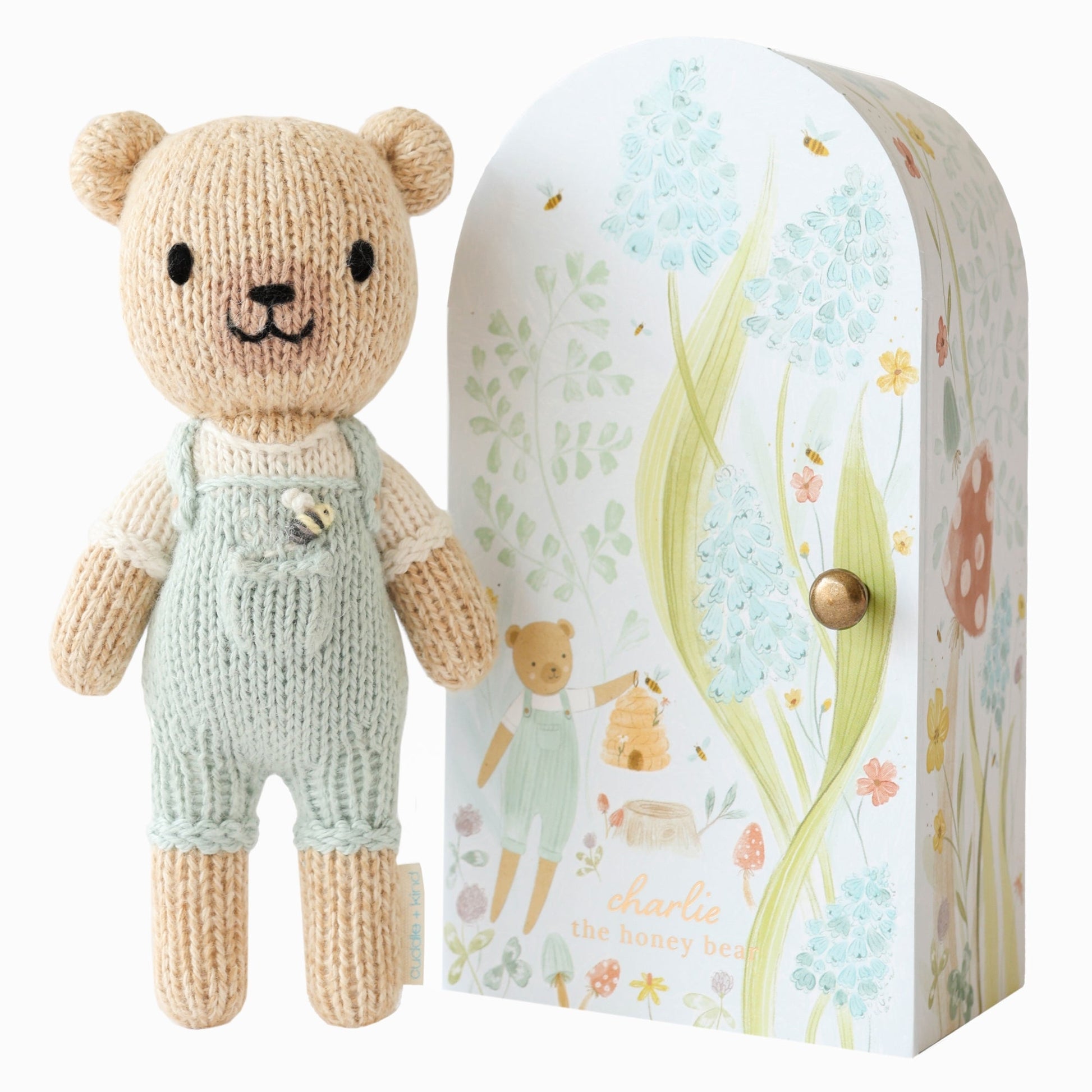 Knitted teddy bear in overalls next to its packaging with floral design. Hand knit doll with premium 100% cotton & floral tiny home. 1 doll provides 10 meals to children in North America and around the world. 