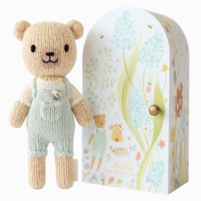 Knitted teddy bear in overalls next to its packaging with floral design. Hand knit doll with premium 100% cotton & floral tiny home. 1 doll provides 10 meals to children in North America and around the world. 