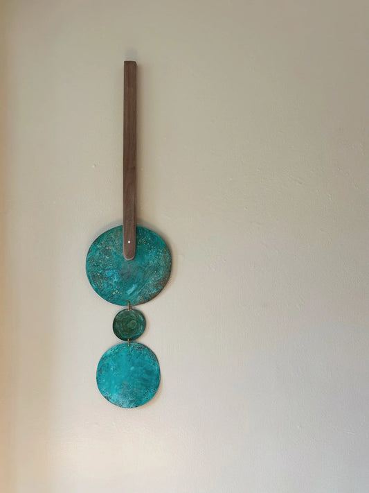 Decorative wall hanging with three turquoise circular discs and a wooden stick on a plain background. Wall hanging made of patinated brass pieces and hammered brass rod connection and a wooden shape. Handmade in NorCal.