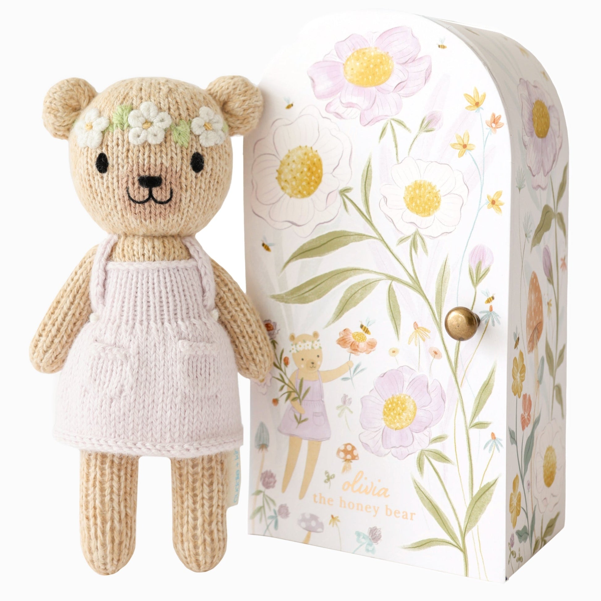 Knitted teddy bear with a floral headband next to a decorative box with floral patterns. Hand knit doll with premium 100% cotton & floral tiny home. 1 doll provides 10 meals to children in North America and around the world. 