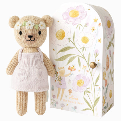 Knitted teddy bear with a floral headband next to a decorative box with floral patterns. Hand knit doll with premium 100% cotton & floral tiny home. 1 doll provides 10 meals to children in North America and around the world. 