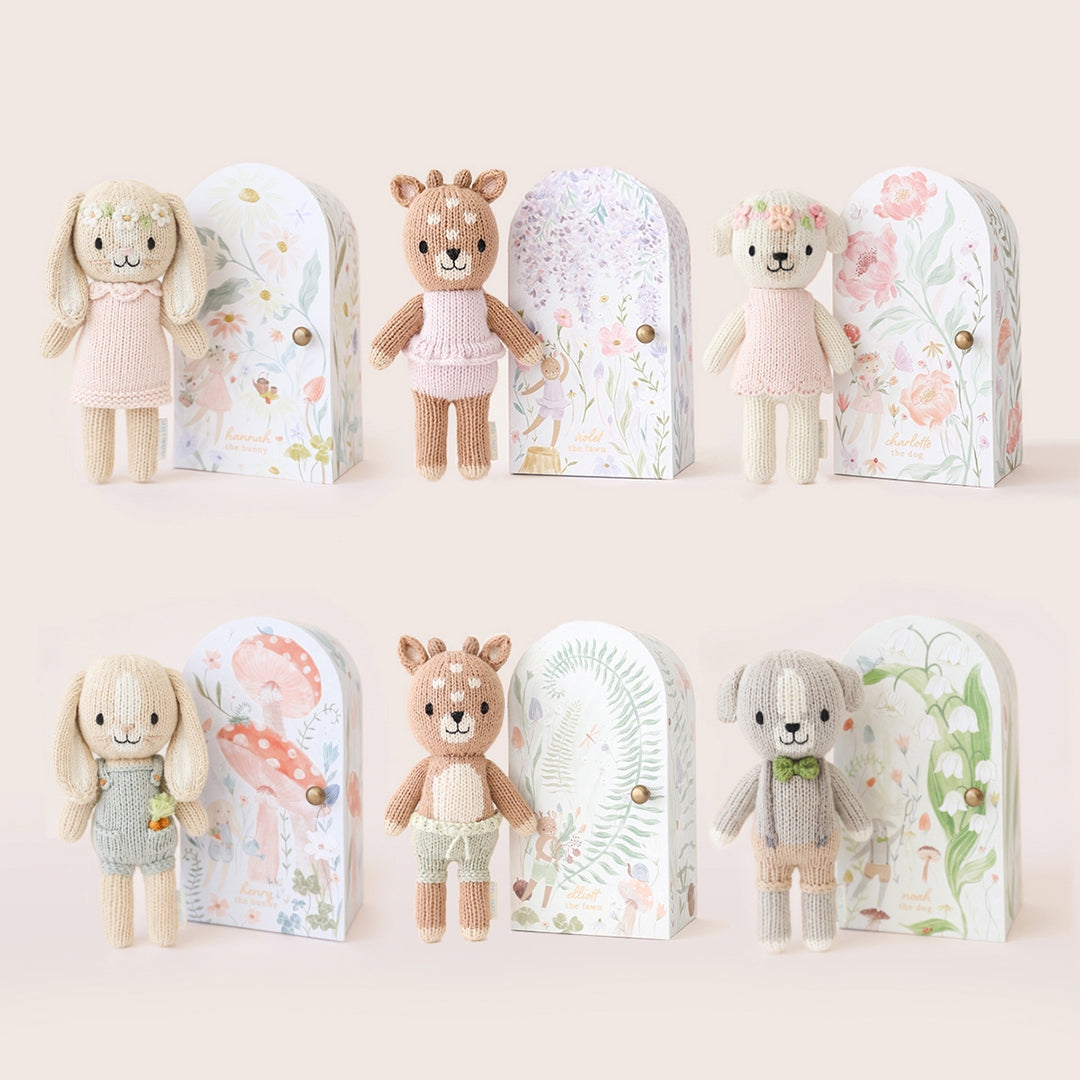Set of stuffed animals in various outfits with decorative boxes on a light background. Hand knit doll with premium 100% cotton & floral tiny home. 1 doll provides 10 meals to children in North America and around the world. 