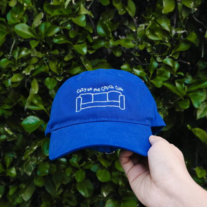 Blue cap with a couch design held against a green leafy background Cozy On the Couch Club Embroidered Hat
