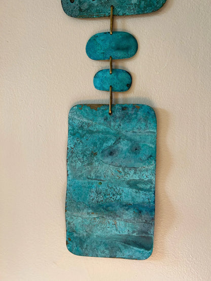 Turquoise stone pendant on a beige background. Wall hanging made of patinated brass pieces and hammered brass rod connection and a wooden shape. Handmade in NorCal.