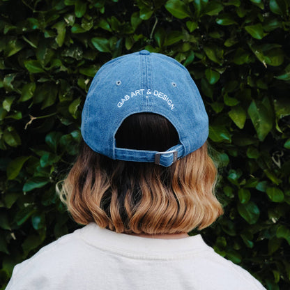 Person wearing a blue cap with 'GAR ART & DESIGN' text against a green hedge background Artsy Embroidered Hat 