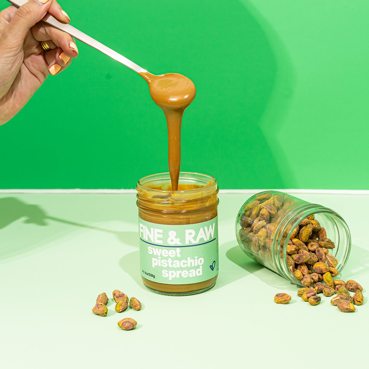 Hand holding a spoon with sweet pistachio spread over a jar of the spread and pistachios on a green background. fine and raw sweet pistachio spread