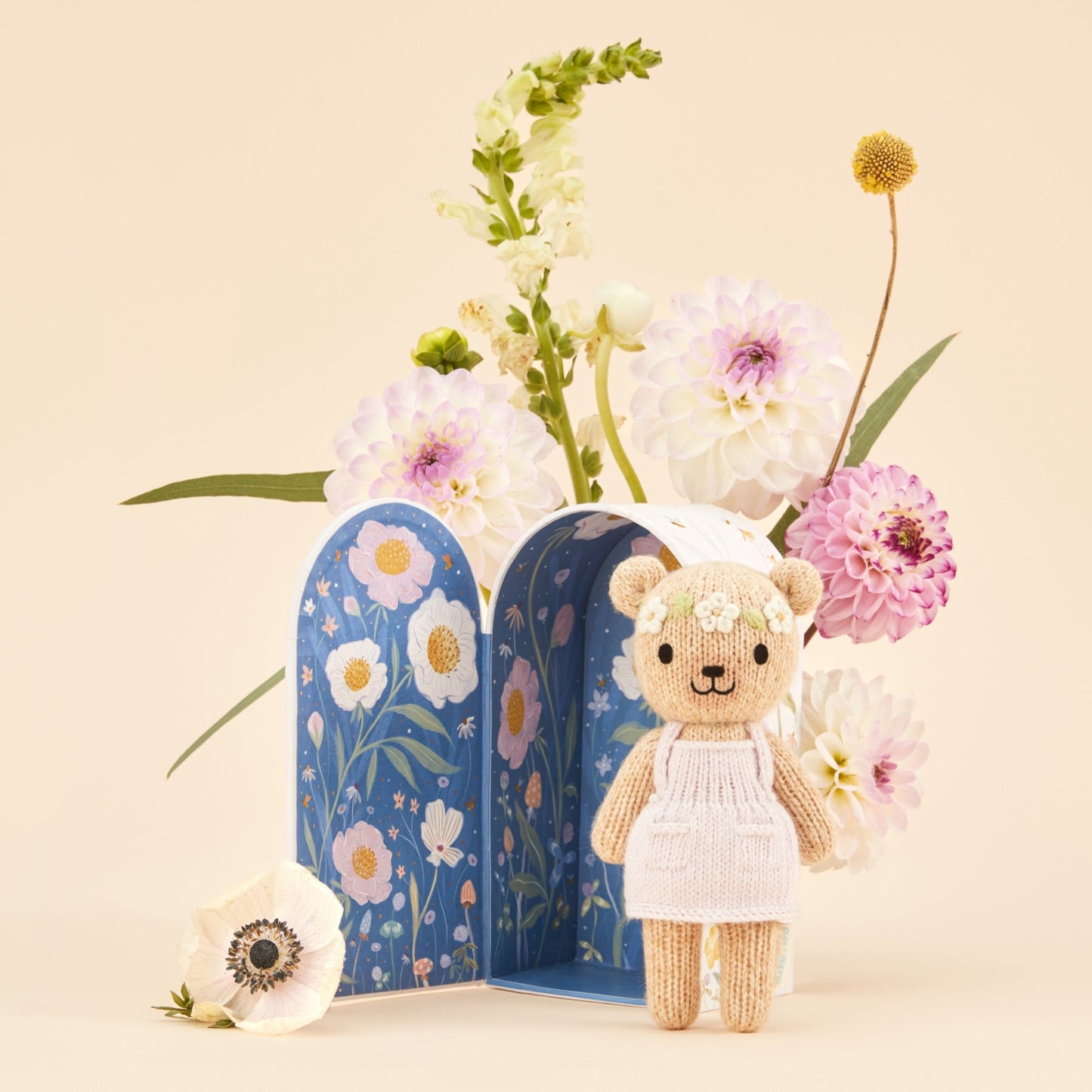 Teddy bear in a white dress standing next to floral bookmarks and flowers on a beige background. Hand knit doll with premium 100% cotton & floral tiny home. 1 doll provides 10 meals to children in North America and around the world. 
