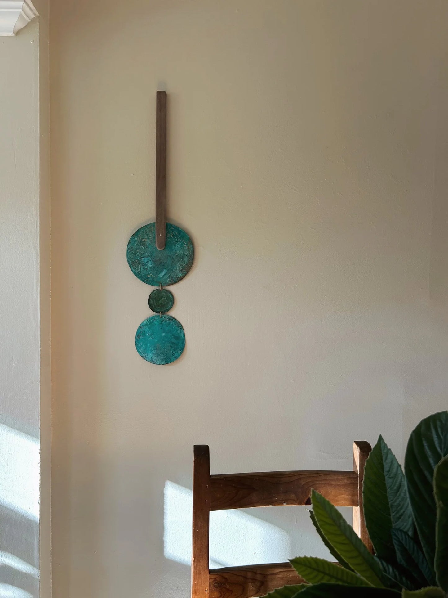 Decorative wall art with circular designs on a beige wall, with a wooden chair and plant in the foreground. Wall hanging made of patinated brass pieces and hammered brass rod connection and a wooden shape. Handmade in NorCal.