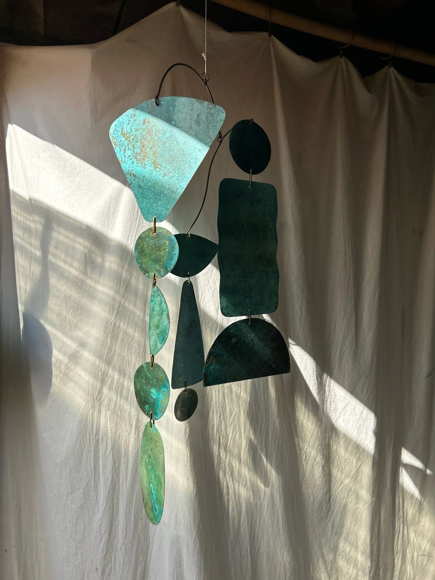 Decorative hanging mobile with teal and green shapes against a white curtain. Kinetic piece made of patinated brass pieces and manipulated brass rod. Handmade in NorCal.