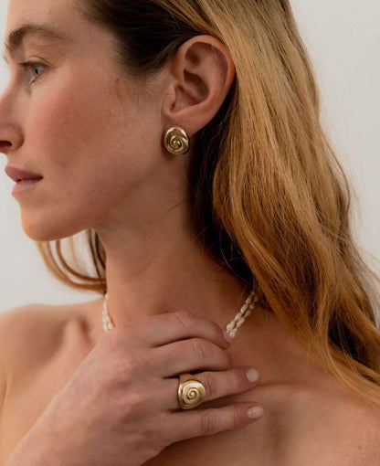 Woman wearing gold spiral earrings and a ring, with a plain background. Amanda Hunt Compass Earrings