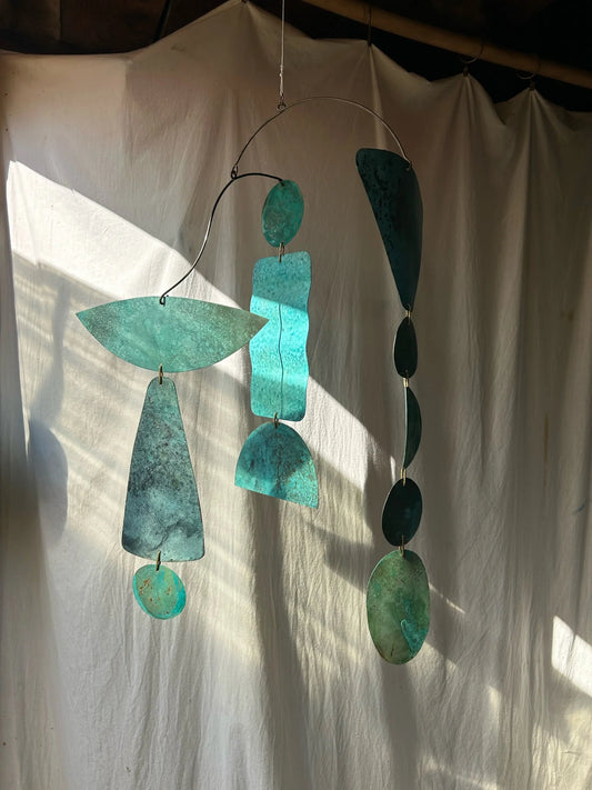 Turquoise metal mobile hanging against a white curtain. Kinetic piece made of patinated brass pieces and manipulated brass rod. Handmade in NorCal.