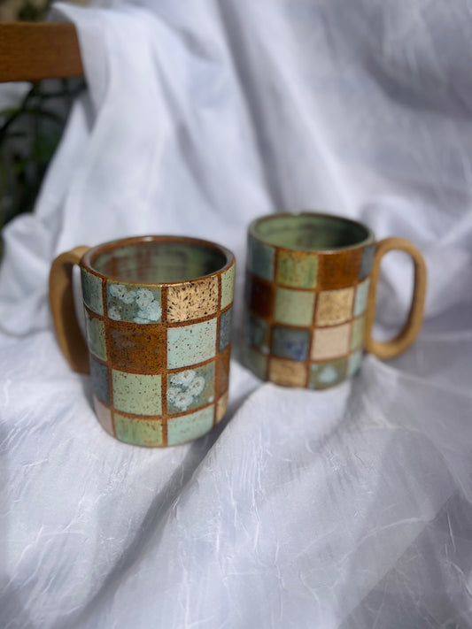 Two ceramic mugs with checkered pattern on a white fabric background