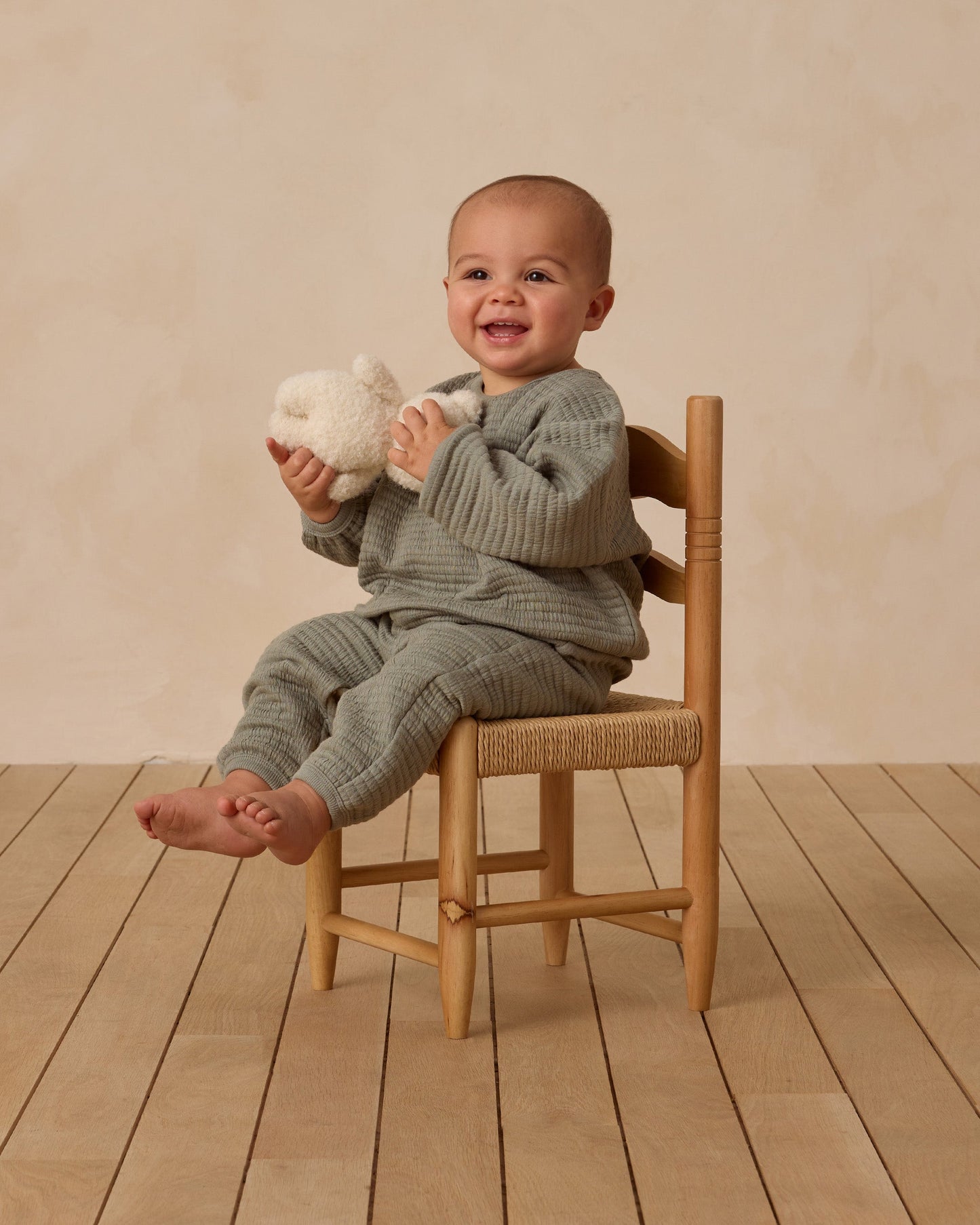 Child sitting on a wooden chair holding a plush toy against a beige background. quincy mae textured sweat set