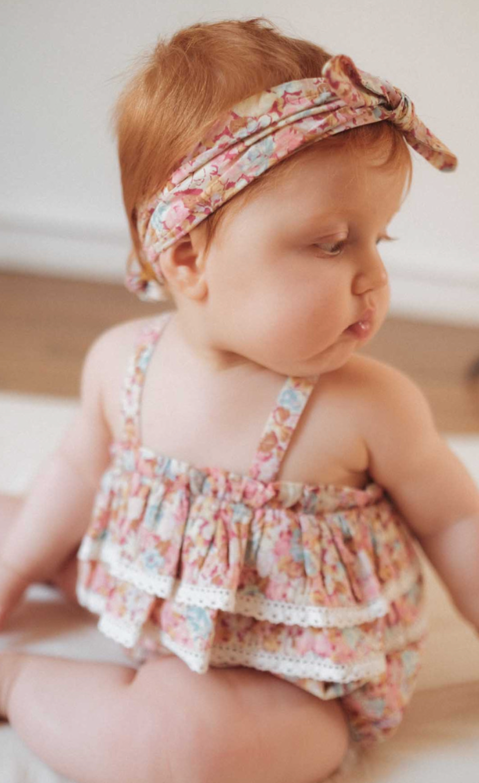 Louise Misha Kumal Romper in sweet pink pastel. A floral print romper adorned with lace edged ruffles that features adjustable, elasticized, straps crossed in the back, elasticized legs, and a crotch snap opening. Made with 100% organic cotton including the lining.