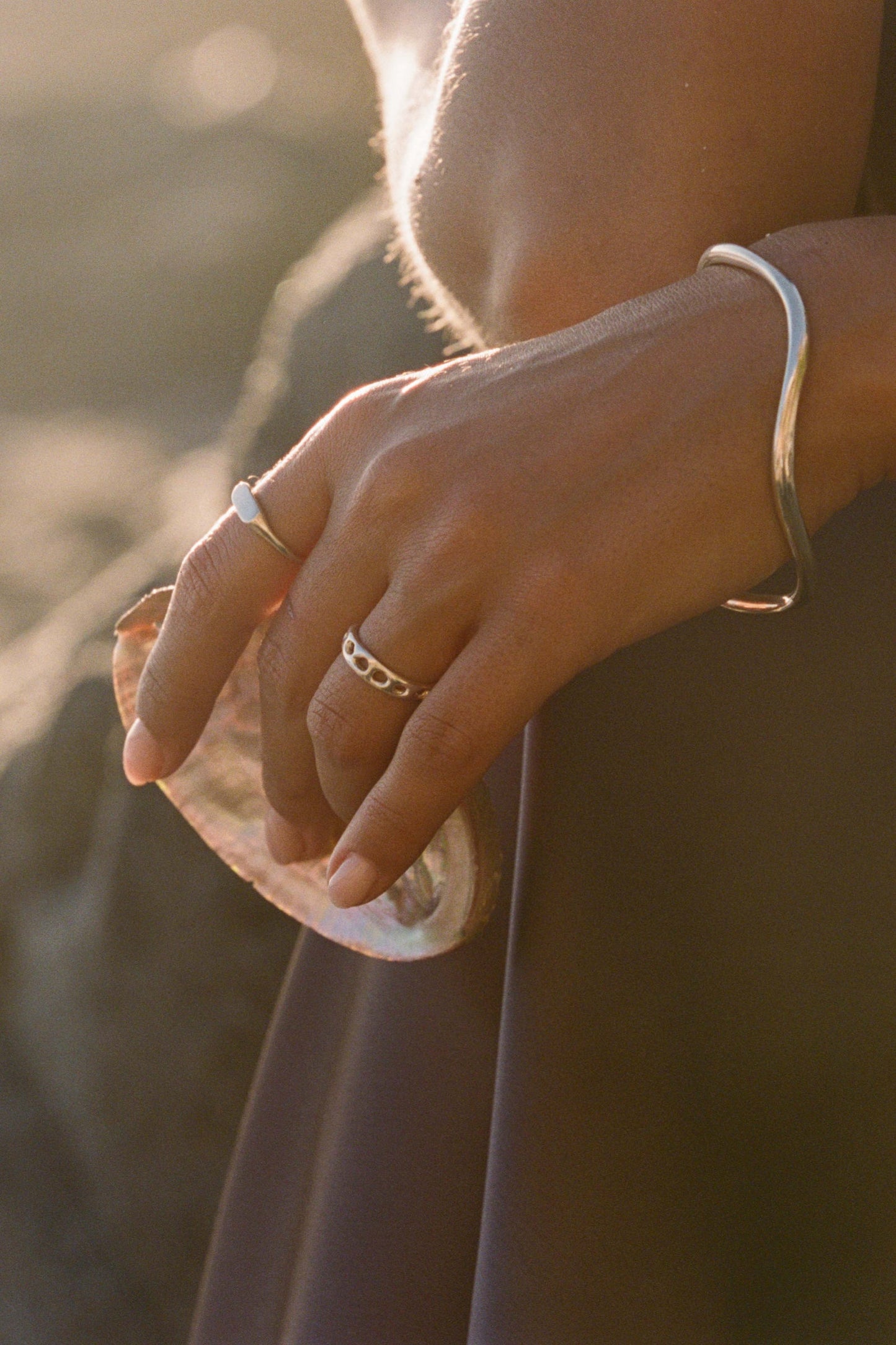 Close-up of a person's hand holding a shell with sunlight casting a warm glow on her handmade sterling silver Moneh Brisel jewelry.