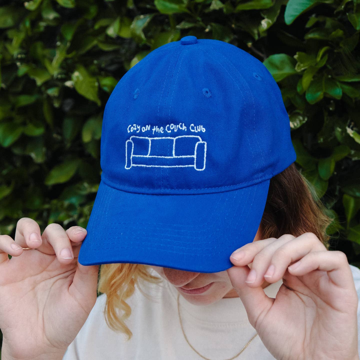 Person holding a blue cap with a logo and text against a green leafy background Cozy On the Couch Club Embroidered Hat