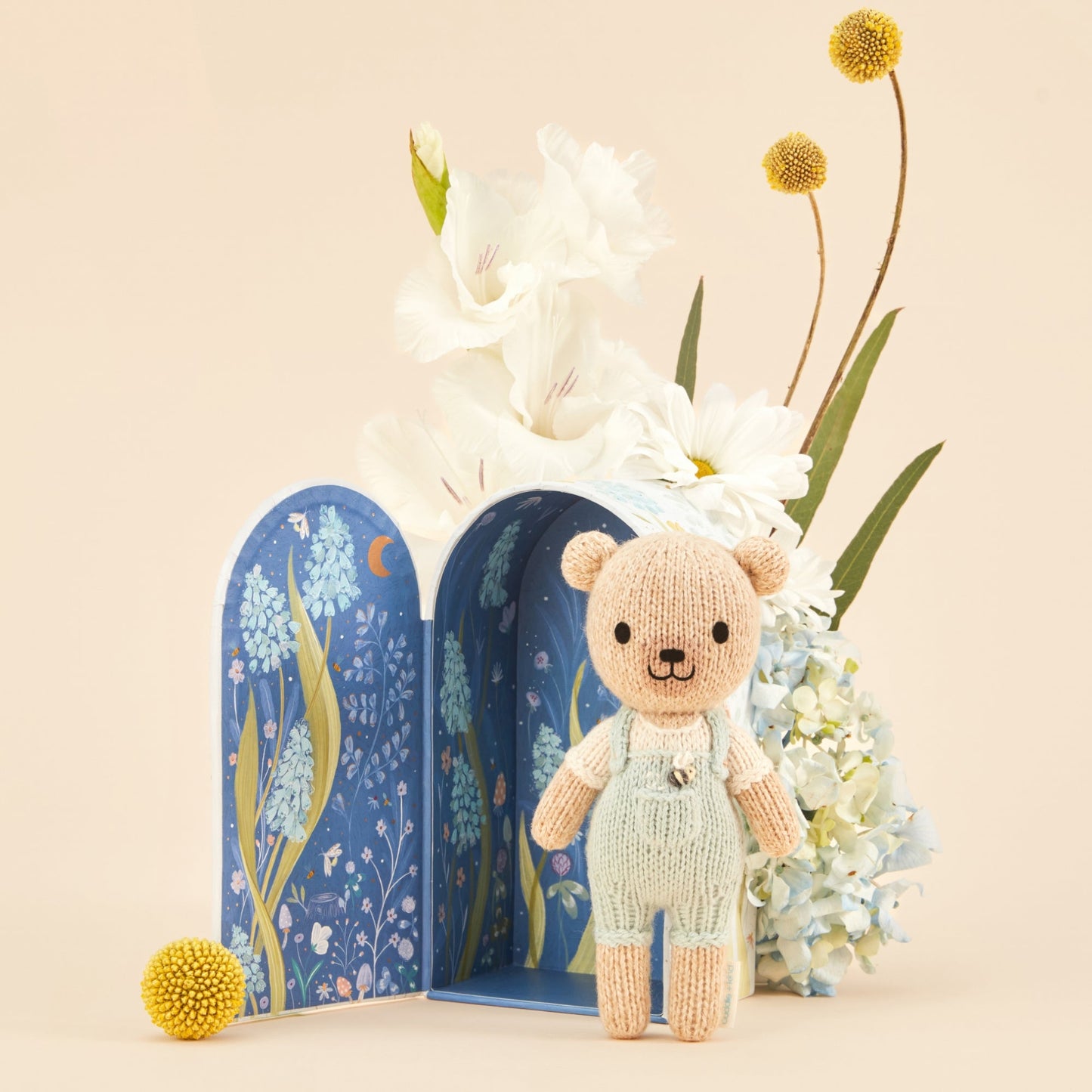 Knitted bear toy in front of a decorative book with floral illustrations and flowers on a beige background. Hand knit doll with premium 100% cotton & floral tiny home. 1 doll provides 10 meals to children in North America and around the world. 