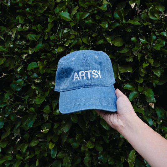 Blue cap with 'ARTSY' text held against a green leafy background Artsy Embroidered Hat 
