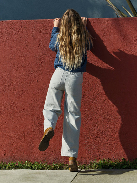 Person with long hair standing against a red wall, wearing a blue sweater and light-colored pants. Mollusk Surf Shop Mid Rise Painter Pants 