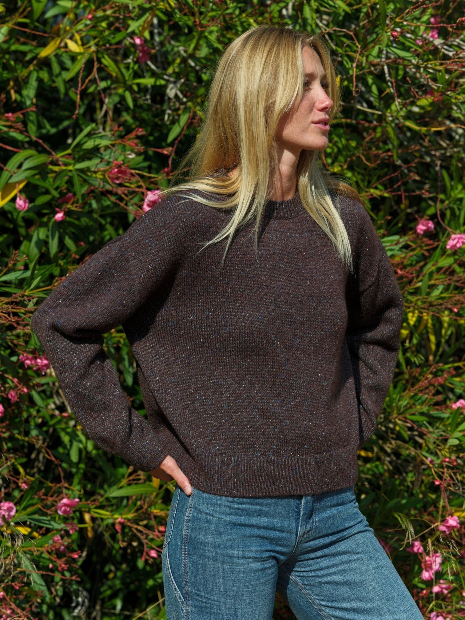Woman wearing a brown sweater and blue jeans standing in front of green foliage with pink flowers. Mollusk Surf Shop Rhys Sweater
