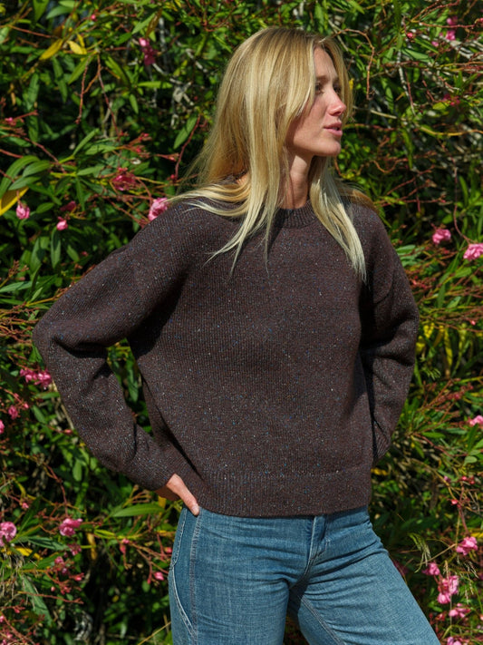 Woman wearing a brown sweater and blue jeans standing in front of green foliage with pink flowers. Mollusk Surf Shop Rhys Sweater