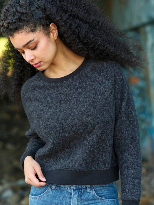 Person wearing a dark gray sweater and blue jeans against a blurred background. Mollusk Surf Shop Softest Crew
