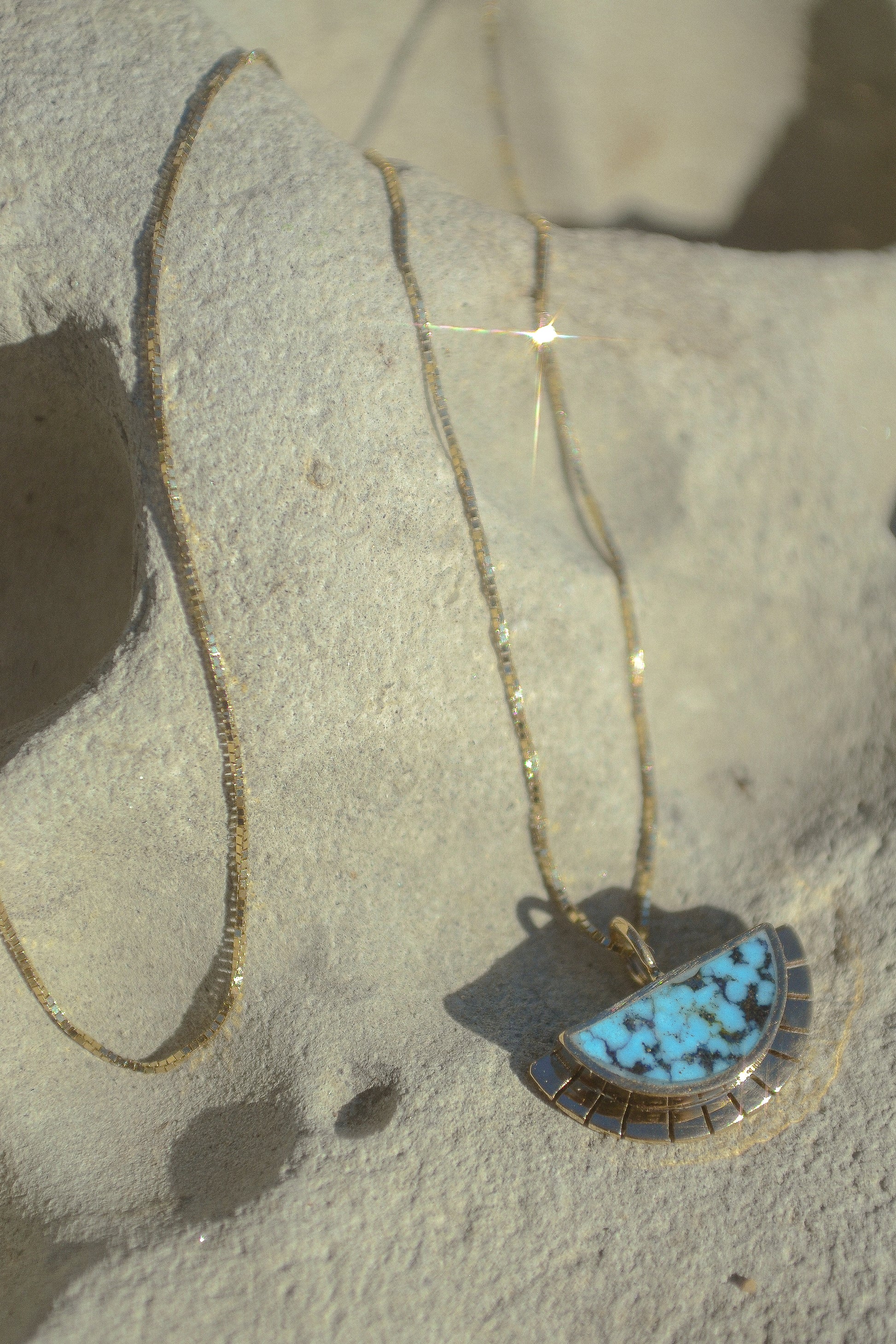 A half circle of warm 14k yellow gold is wrapped around an inlaid Kingman turquoise stone while a serrated halo of light radiates from this sun. This pendant hangs freely on a 14k yellow gold box chain. Handmade in the Rocky Mountains.